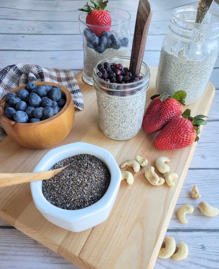 jars and a glass of dairy-free chia pudding topped with blueberries and strawberries. Cashew nuts on the side.