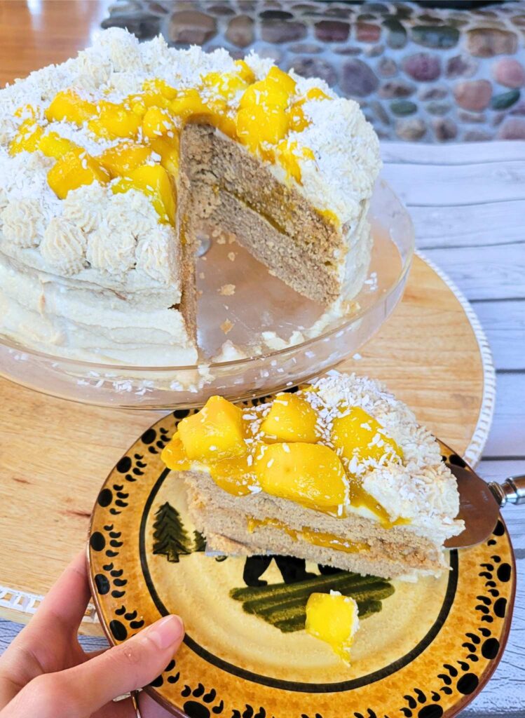 The Gluten-free Mango Cake with dairy-free coconut cream frosting on a platter. A slice of the cake removed and on a plate. 