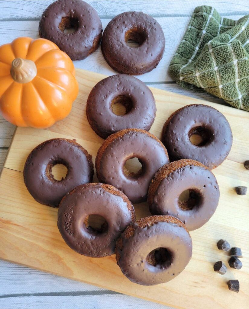 Gluten-free pumpkin donuts with a dark chocolate glaze laying on a cutting board next to a fake pumpkin