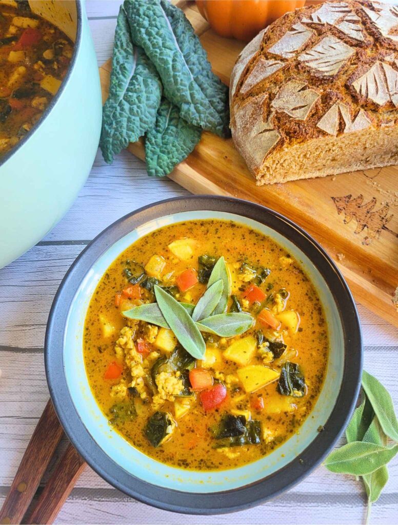 bowl of gut-healing pumpkin chicken soup with fresh sage garnish next to a loaf of einkorn sourdough and fresh kale.